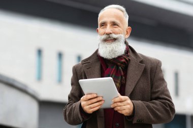 Uzun gri sakallı neşeli yaşlı adam sokakta yürürken dijital tablet tutuyor fotokopi alanına bakıyor ve gülümsüyor. Üst düzey işadamı şık giyinmiş, evi ofis binasının yanında duruyor.