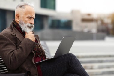 Düşünceli kıdemli sakallı iş adamı dizüstü bilgisayarı ile bankta oturuyor, klavyede daktilo yazıyor, e-posta yolluyor, boşluk yaratıyor. Tarz sahibi, yaşlı, gri saçlı bir adam caddede iş merkezinde oturuyor, internette çalışıyor.
