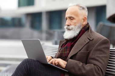 Yoğun gri saçlı, uzun sakallı, sıcak ve şık giyimli bir adam ofis binasının yanındaki bankta oturuyor, dizüstü bilgisayar kullanıyor, internette çalışıyor, müşterilerle sohbet ediyor ya da web sörfü yapıyor.