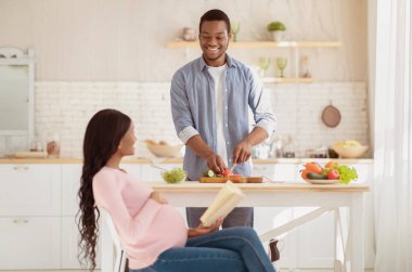 Mutlu siyah adam mutfakta hamile karısına taze sebze salatası yapıyor. Kocası evde sağlıklı yemek pişirirken Afro-Amerikan kadın kitap okuyacak.