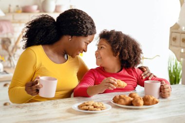 Siyah anneyi ve küçük kızı sevmek, mutfakta atıştırmalık yemek, taze pişmiş kruvasan yemek ve kakao içmek, evde kaynaşırken mutlu Afrika ailesi ev yapımı hamur işi yemek,