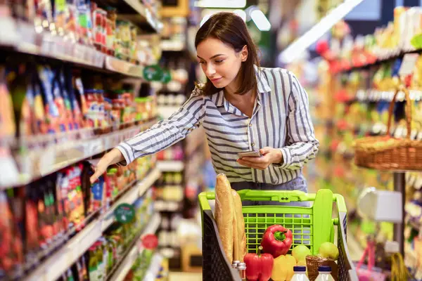 Millennial Lady Holding ve Smartphone 'un portresi. Süpermarkette Trolley Cart' la yemek alışverişi yapıyor. Kadın müşteri listesiyle alışveriş yapıyor, mağazadaki raflardan ürünler alıyor.