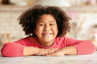 Mutlu Çocuk. Mutfakta masada oturan, Afro-Amerikan bayan çocuğun evde dinlenip, kameraya bakıp gülümseyen, boş alan arayan neşeli siyah kızın yakın plan portresi.