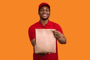 Hızlı Teslimat. Kırmızı şapkalı ve üniformalı Afro-Amerikan kuryenin restorandan kameraya, sarı turuncu stüdyo arka planında el işi kağıt torba taşıdığı ve verdiği ön manzara.