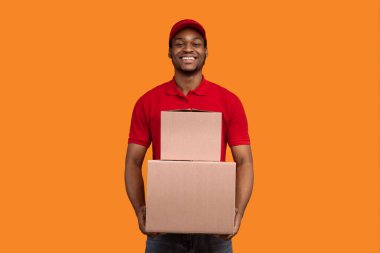 Teslimat servisi konsepti. Kırmızı şapkalı ve üniformalı gülümseyen Afro-Amerikan erkek kurye elinde karton kutular, paketlerle turuncu stüdyo arka planında tek başına duruyor.