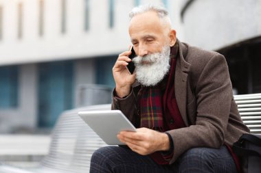 Uzun sakallı yaşlı bir adam ofis binasının yanındaki bankta oturuyor, dijital tablet ekranına bakıyor ve cep telefonuyla konuşuyor, sokakta dinlenirken iş görüşmesi yapıyor, boş bir alanda.