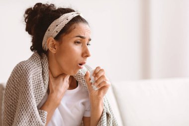 Hasta, genç esmer kadın, battaniyeye sarılmış öksürüyor ve fotokopi odasına bakıyor, oturma odasında kanepede oturuyor, yakın plan. Öksüren genç bayan, koronavirüs ya da viral zatürree.