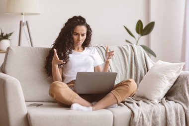 Sinirli kadın serbest çalışan dizüstü bilgisayara ve hareketlere bakıyor, oturma odasının içi, kopyalama alanı. Duygusal genç esmer bayan işiyle ilgili sorunlar yaşıyor, evdeki kanepede oturuyor.