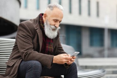 Sokakta dinlenirken akıllı telefon kullanan, bankta oturan ve mobil uygulama kullanan mutlu gri saçlı adam, kopyalama alanı. Dışarıda akıllı telefon kullanan yakışıklı bir son sınıf öğrencisi.
