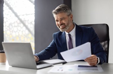 Successful middle aged entrepreneur working with laptop and papers at workplace in modern office, smiling mature businessman in suit sitting at desk and managing financial reports, copy space