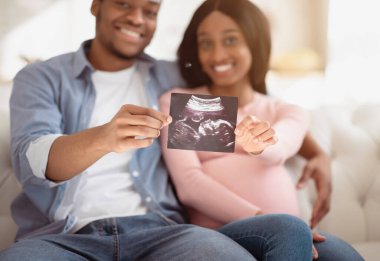Geleceğin genç ebeveynleri ellerinde doğmamış bebeklerinin resimlerini tutuyorlar. Ellerine odaklanmış durumdalar. Çocuğunun sonogramıyla birlikte mutlu anılar yaratan pozitif beklenen bir çift.