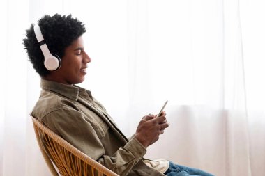 Kulaklıklı Afro-Amerikalı genç adam evde sandalyesinde otururken akıllı telefondan müzik dinliyor, boşluğu kopyalıyor. Koltuğunda dinlenen siyah bir gencin yan görüntüsü çevrimiçi radyo yayınının keyfini çıkarıyor.