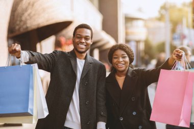 Mevsimlik satışlarda ve tatilden önce Kara Cuma 'da çok alışveriş. Paltolu mutlu Afro-Amerikan erkek ve kadın. Renkli alışveriş torbaları gösteriyor ve kasabanın yakınındaki dükkânda başarılarını dile getiriyor.