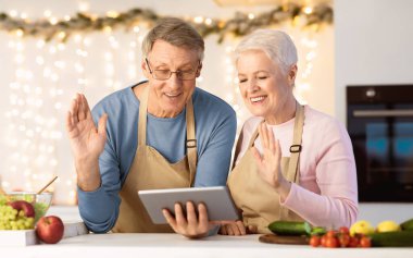 Son sınıf çifti, Noel arifesinde evde mutfakta dikilirken dijital tablete el sallıyor. Modern Mesafe Aile İletişim ve Xmas Kutlaması Çevrimiçi