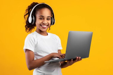 Çocuklar ve Teknoloji Konsepti. Gülümseyen siyah kızın portresi dizüstü bilgisayar kullanıyor, kablosuz kulaklık takıyor, çalışıyor, çevrimiçi kurslar izliyor, sarı stüdyo duvarından izole edilmiş.