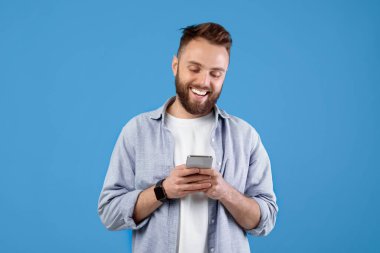 Mavi stüdyo arka planında akıllı telefon kullanan beyaz bir gencin portresi. Modern aletlerle gülümseyen milenyum adamı internette sohbet ediyor, internette geziniyor, sosyal medyada paylaşıyor.