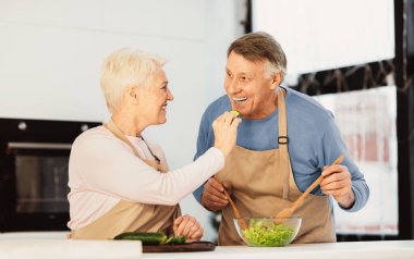Mutlu son sınıf çifti birbirlerini besliyor ve gülüyor modern mutfakta birlikte yemek pişiriyorlar. Yaşlı eşler zevkli yemekler tadıyor. Aile Yemeği Hazırlama