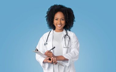 Gülümseyen Afro-Amerikan kıvırcık kadın doktor beyaz önlüklü terapist tablete not alıyor mavi arka planda, stüdyoda. Sağlık hizmeti ve muayene, sağlık ve tedavi