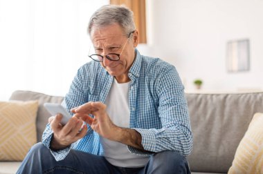Zavallı görme duyusu, yaşlılık çağı, Maküler Bozukluk Konsepti. Gözlüklü, kafası karışmış, şaşı, şaşı gözlü, cep telefonunda mesaj okuyan, sehpada otururken görme sorunu yaşayan yetişkin bir erkek.