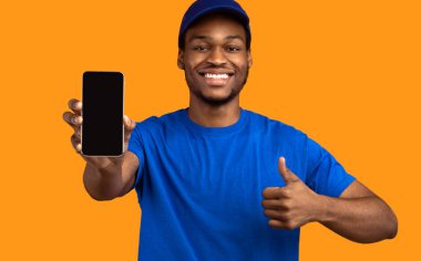 Teslimat Servisi öneriliyor. Gülümseyen Afro-Amerikan erkek kurye elinde boş ekranlı akıllı telefon ve mavi şapkalı, üniformalı, turuncu renkli bir stüdyo.