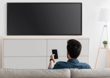 Modern aletler. Akıllı telefonu TV 'ye bağlı, evde video izleyen, arka görüşlü tanınmayan bir adam. Adam telefondan televizyona video aktarıyor. Kablosuz ağ üzerinden aygıtlar arasındaki bilgi aktarımı.