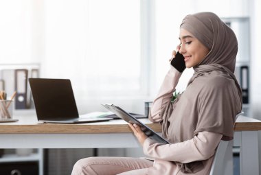 Başörtüsü tutan Müslüman iş kadını, belgeyi kontrol ediyor ve iş yerinde otururken ortağıyla cep telefonundan konuşuyor.