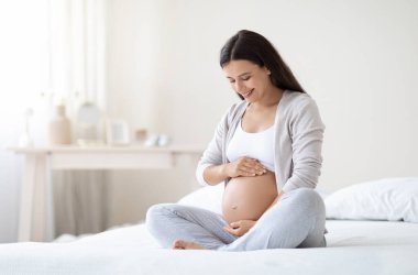 Happy pregnant woman sitting on bed in bedroom at home, hugging her big tummy. Happiness and feeling joyful as emotional well affect to baby neurological and psychological development, empty space