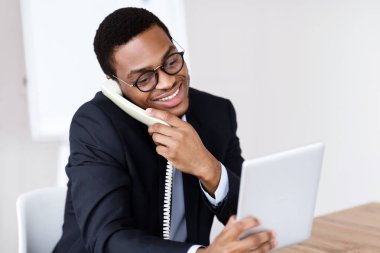 Takım elbiseli mutlu siyah adam ofis yöneticisi veya iş adamı telefonda konuşuyor, çalışma masasında dijital tablet kullanıyor, müşterilere danışıyor, modern teknolojiler kullanıyor, kopyalama alanı, ofis içi