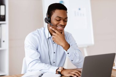 Pozitif genç siyahi adam ofiste online iş konferansı yapıyor, dizüstü bilgisayar ve kulaklık kullanıyor, müşteri ya da iş ortağıyla neşeli bir şekilde konuşuyor, bilgisayar ekranına bakıyor ve gülümsüyor, boşluğu kopyalıyor.