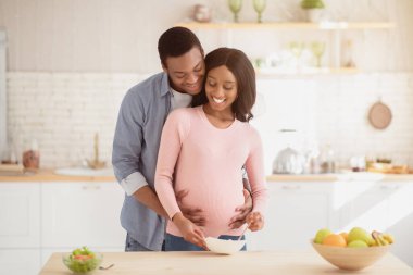Güzel siyah hamile kadın mutfakta sağlıklı kahvaltı hazırlıyor ve kocası ona yardım ediyor. Bekleyen Afrikalı Amerikalı çift dengeli yemek pişiriyor, içeride birlikte vakit geçiriyorlar.