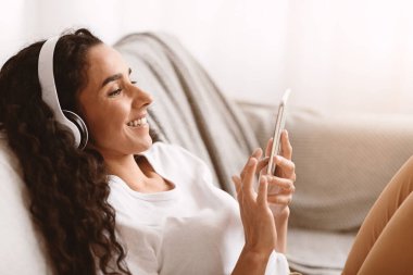 Uzun kıvırcık saçlı, uzun kıvırcık saçlı, film izleyen veya cep telefonuyla komik videolar izleyen, cep telefonu uygulaması kullanan, evdeki koltukta oturan mutlu esmer kadın.