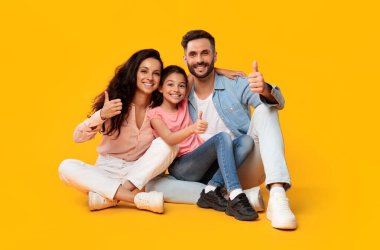 Üç kişilik mutlu bir aile, bir anne, bir baba ve küçük bir kız, sarı bir arka planda oturuyor, gülümsüyor ve başparmak işareti yapıyor. Aile gündelik giysiler içinde, herkes kameraya bakıyor..