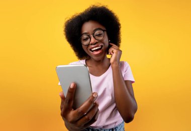 Mutlu Afro-Amerikan bayan akıllı telefon kullanıyor ve video görüşmesi yapıyor, cep telefonu kamerasına gülümsüyor, sarı arka planda poz veriyor. Heyecanlı zenci kadın telefonda selfie çekiyor.