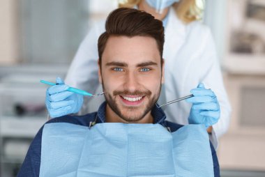 Gülümseyen genç sakallı adam, diş hekimliği koltuğunda oturuyor, modern diş kliniğini ziyaret ediyor, düzenli sağlık kontrolü yaptırıyor, diş hekimi tanınmayan bir kadın diş hekimi, erkek hasta ağzının yanında diş aleti tutuyor.