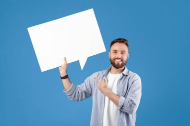 Mutlu genç adam boş konuşma balonunu işaret ederek reklam ya da tanıtım için mavi stüdyo arka planına bakıyor, maket. Beyaz bulutlu yakışıklı beyaz adam duyuru yapıyor.