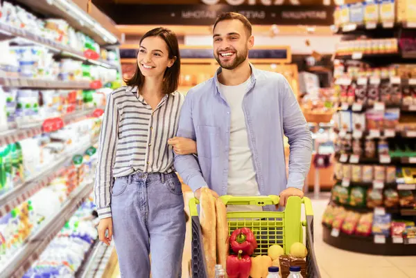 Sağlıklı beslenme. Markette alışveriş yapan mutlu ve genç bir çift, neşeli müşteriler kapalı alanda mandıralı rafların yanında Trolley Cart ile gezip yiyecek seçiyor ve satın alıyorlar.