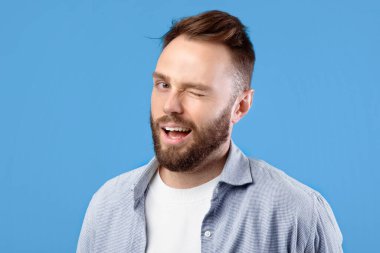 Kendine güvenen yakışıklı beyaz adam mavi stüdyo arka planında kameraya göz kırpıyor ve gülümsüyor. Sıradan giyinmiş, oynak, genç sakallı adam flört ediyor, arkadaş canlısı tavırlar sergiliyor.