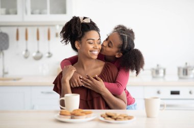 Sevimli siyah kız kahvaltıdan sonra güzel annesine sarılıp öpüyor. Mutfağın içinde. Afro-amerikan anne-kız kaynaşırken abur cubur yemek, kopyalama alanı