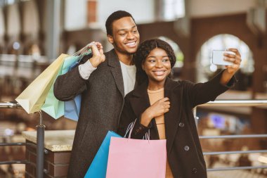 Sezon indirimi sırasında ve Kara Cuma 'da birlikte alışveriş yapmak. Mutlu yıllar Afro-Amerikan modasına uygun kadın ve montlu adam alışveriş merkezinde yürürken akıllı telefondan selfie çekiyor.