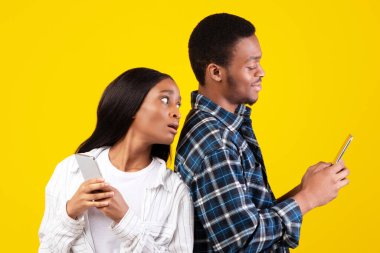 Kıskançlık, internet ağları ve ilişkiler. Bin yıllık Afro-Amerikan öğrenci bayan gülümseyen kocanın telefonuna bakar, sarı arka planda izole edilmiş, boş alan, stüdyo çekimi.