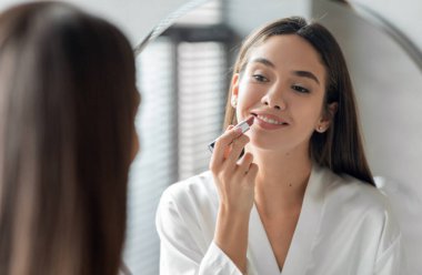 Makyaj rutini. Güzel genç bir kadın banyoda aynanın karşısında dikilirken dudaklarına çıplak ruj sürüyor, çekici bir kadın yansımasına gülümsüyor, seçici bir şekilde odaklanıyor.