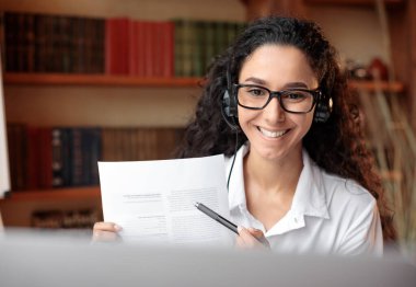 Uzaktan kumandalı çalışma. Gözlüklü ve kulaklıklı gülümseyen genç bir kadının portresi bilgisayarını kullanarak, kağıt tutarak, rapor tutarak, sözleşme belgelerini ya da web kamerasına anlaşmalarını göstererek sanal görüntüler elde ediyor.