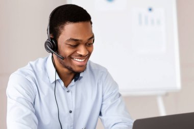 Keyifli siyahi adam kriz yöneticisi ya da müşteri hizmetleri temsilcisi ofiste dizüstü bilgisayar başında çalışıyor, kulaklık takıyor, müşterilerle veya iş ortaklarıyla çevrimiçi sohbet ediyor, kopyalama alanı kullanıyor.