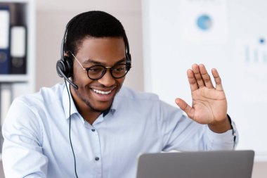 Neşeli siyahi adam iş ortaklarını selamlıyor, ofisten online konferans veriyor, dizüstü bilgisayar ve kulaklık kullanıyor, kopyalama alanı kullanıyor. Genç Afrikalı Amerikalı adam çevrimiçi eğitime katılıyor.
