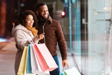 Afro-Amerikan bir çiftin vitrininde bir şey seçerken portresi, seçtiği şeyi işaret eden kadın, akşam alışveriş merkezinin yakınında erkek arkadaşı veya kocasıyla duruyor, alışveriş torbaları taşıyorlar.