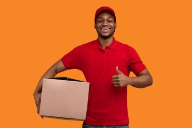 En iyi servis konsepti. Kırmızı şapkalı, üniformalı, elinde karton kutuyla gülümseyen siyah teslimatçı. Sarı turuncu stüdyo arka planında izole bir şekilde duran baş parmağını kaldırıyor.