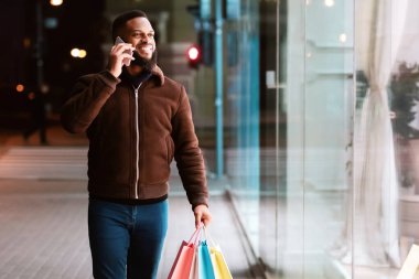 Yaşam tarzı kavramı. Cep telefonuyla konuşan, elinde alışveriş poşetleri olan, akşamları sokakta yürüyen ve vitrinlere bakan gülümseyen bir zenci. Modern İletişim
