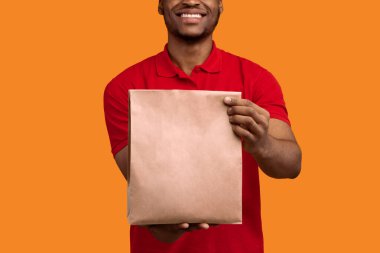 Kırmızı üniformalı Afro-Amerikan teslimatçının, restorandan kameraya ürünlerle dolu el çantası taşıyarak turuncu stüdyo arka planında izole edilmiş,
