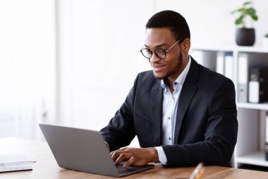 Resmi takım elbiseli neşeli genç siyahi girişimci dizüstü bilgisayarla çalışıyor, gözlük takıyor, ofis içi, fotokopi odası. Başarılı bin yıllık Afro-Amerikan iş adamı iş projesi üzerinde çalışıyor.