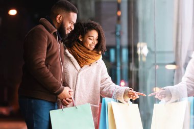 Buraya bak. Güzel Afro-Amerikalı kadın mağaza vitrininde erkeğine moda kıyafetlerini gösteriyor, akşamları alışveriş merkezinin yakınında yürüyor. Kış Satış ve İndirim, Perakende, Satın Alım